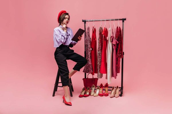 Girl in stylish outfit and red beret sits on chair and thoughtfully reads on pink background with shiny clothes and shoes