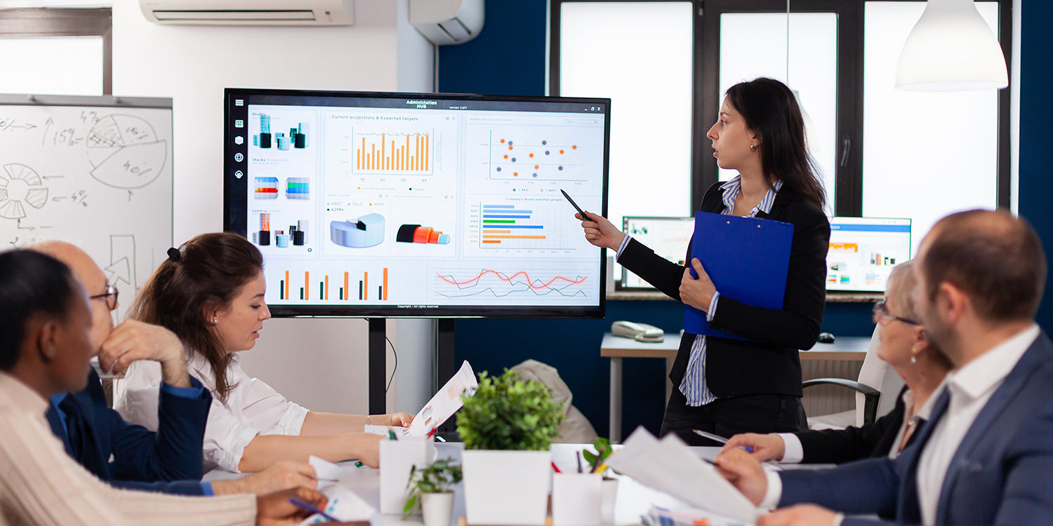 Young team leader in big corporation briefing coworkers pointing at graph meeting corporate staff